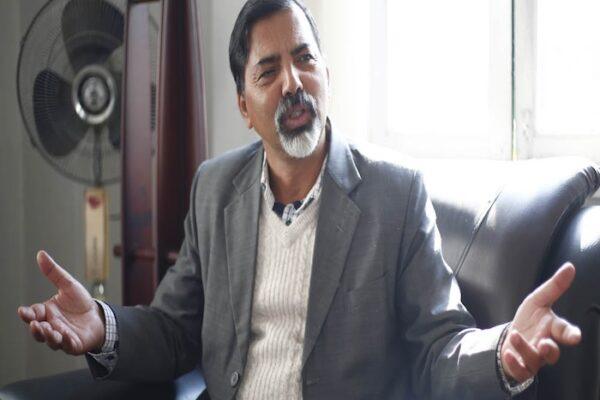 A man in a gray suit and white sweater sitting on a couch, gesturing with his hands while speaking.