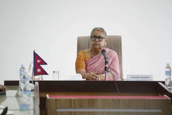 A woman wearing a pink saree seated at a table with a microphone and Nepalese flags in the background.
