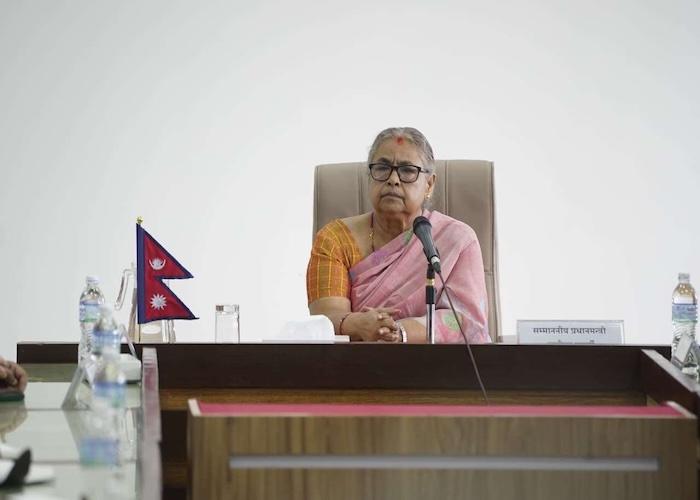 A woman wearing a pink saree seated at a table with a microphone and Nepalese flags in the background.