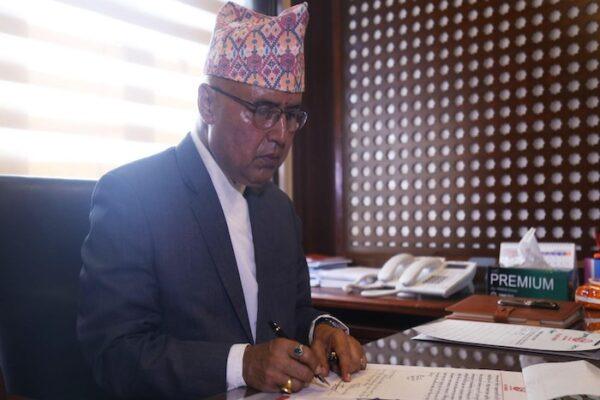 Rameshwor Khanal in a formal suit signing documents at a desk with a patterned background.