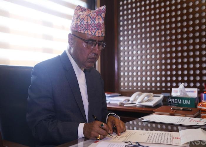 Rameshwor Khanal in a formal suit signing documents at a desk with a patterned background.