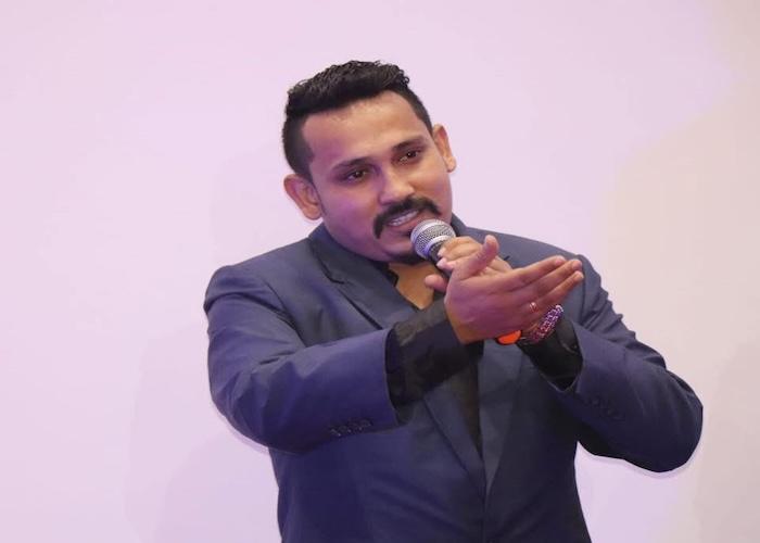 Man in a blue suit holding a microphone while speaking at an event or presentation.