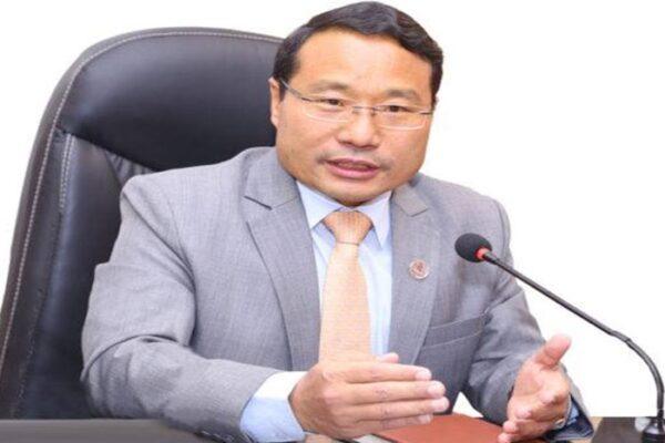 A man in a gray suit with glasses speaking into a microphone while seated at a desk.
