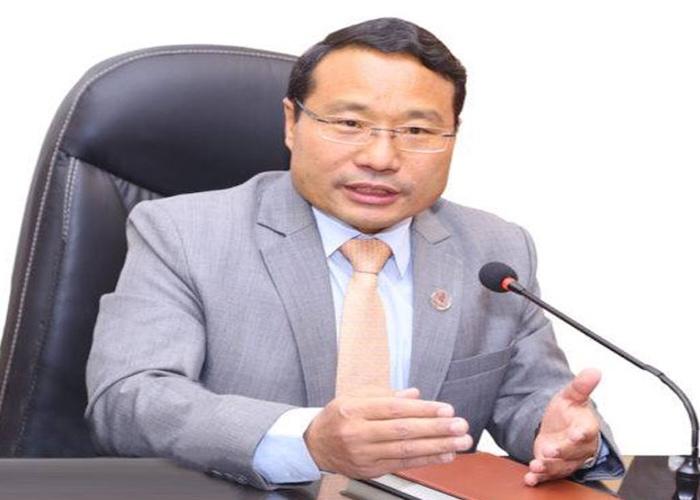 A man in a gray suit with glasses speaking into a microphone while seated at a desk.