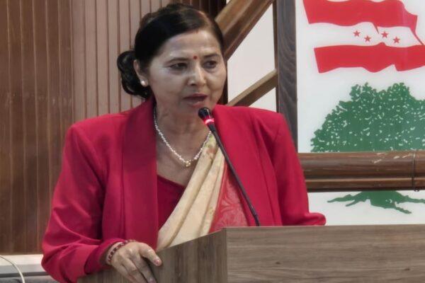 Woman in a red blazer and traditional attire speaking into a microphone at a podium with a national emblem in the background.