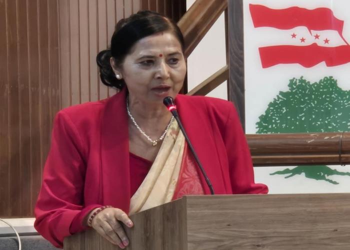 Woman in a red blazer and traditional attire speaking into a microphone at a podium with a national emblem in the background.