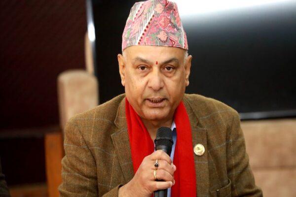 Man wearing a traditional Nepali topi and a red scarf, speaking into a microphone at an event.