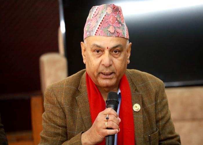Man wearing a traditional Nepali topi and a red scarf, speaking into a microphone at an event.